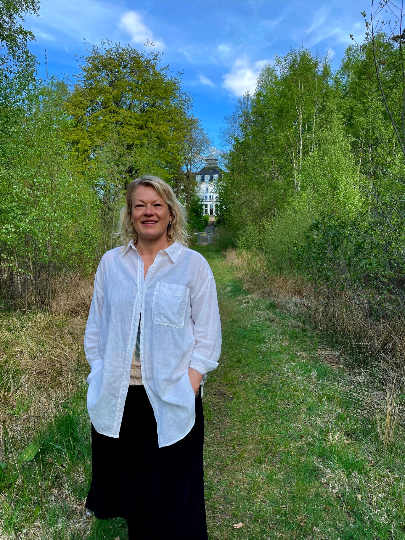 Trine går i skoven med hvidt hus i baggrunden